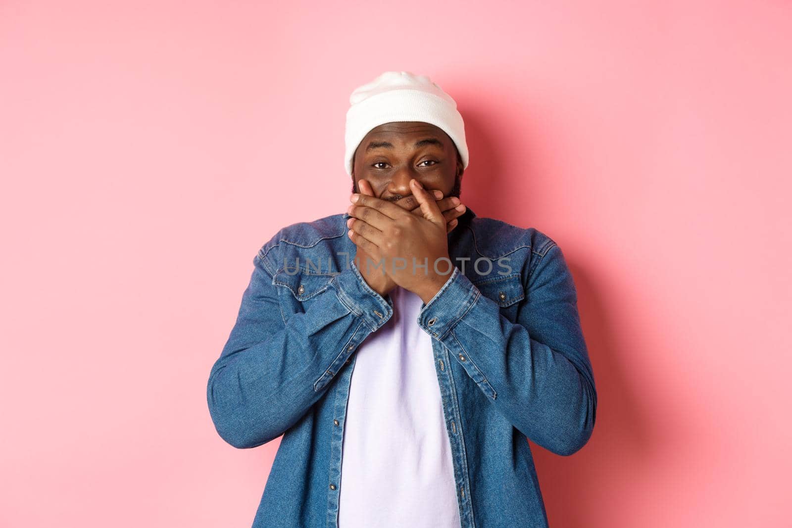 Happy Black hipster man holding laugh, cover mouth and giggle over funny joke, staring at camera and chuckling, standing over pink background.