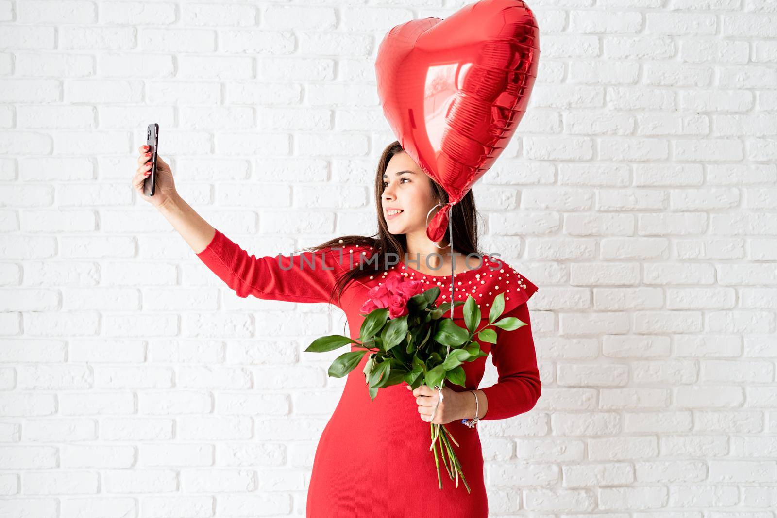 Valentines Day. Young brunette woman in red dress holding a red heart balloon and flowers over white brick wall background