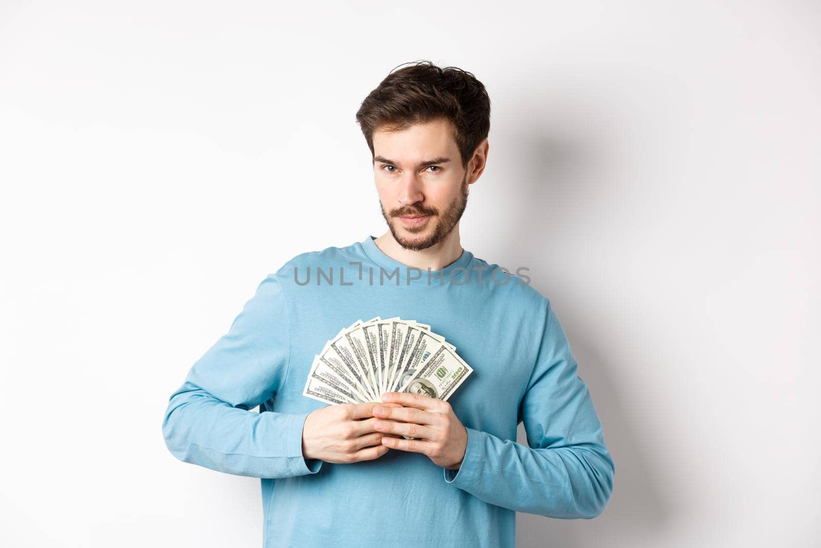 Confident bearded man smiling, showing dollars and looking at camera. Handsome guy holding money on white background.