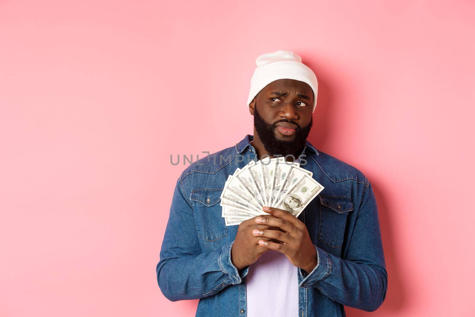 Hesitant african-american man holding money, looking left with doubts and concerns, standing against pink background.