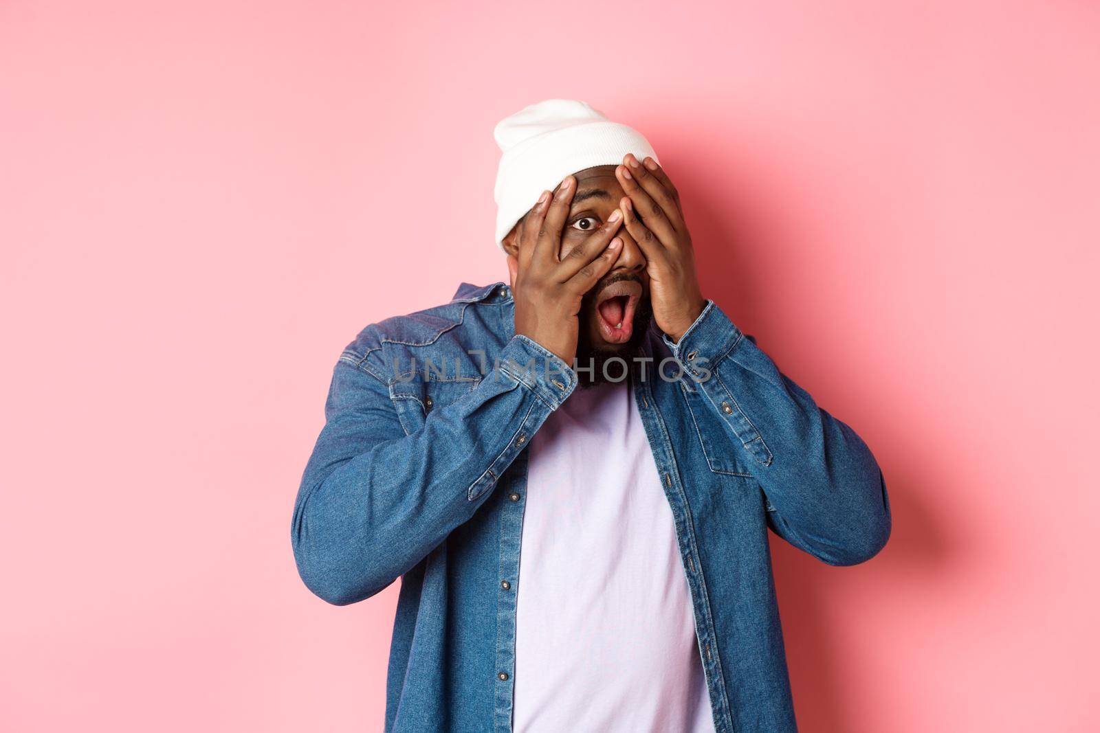 Curious Black man cover eyes but peeking through fingers, staring at camera amazed, standing in hipster beanie against pink background.