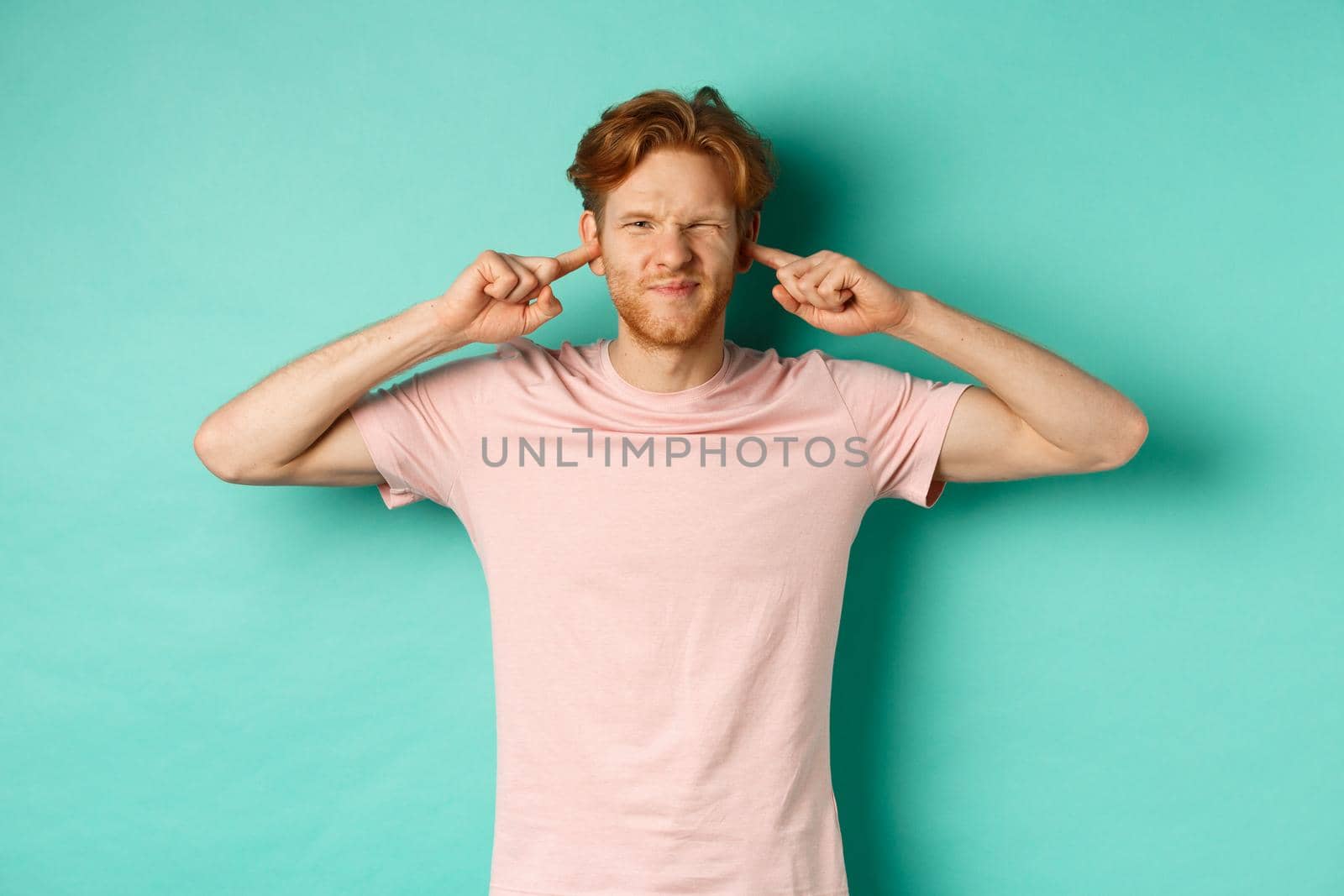 Annoyed young man with red hair and beard shut his ears and grimacing, disturbed by loud bothering sound, noisy neighbours, standing over turquoise background by Benzoix