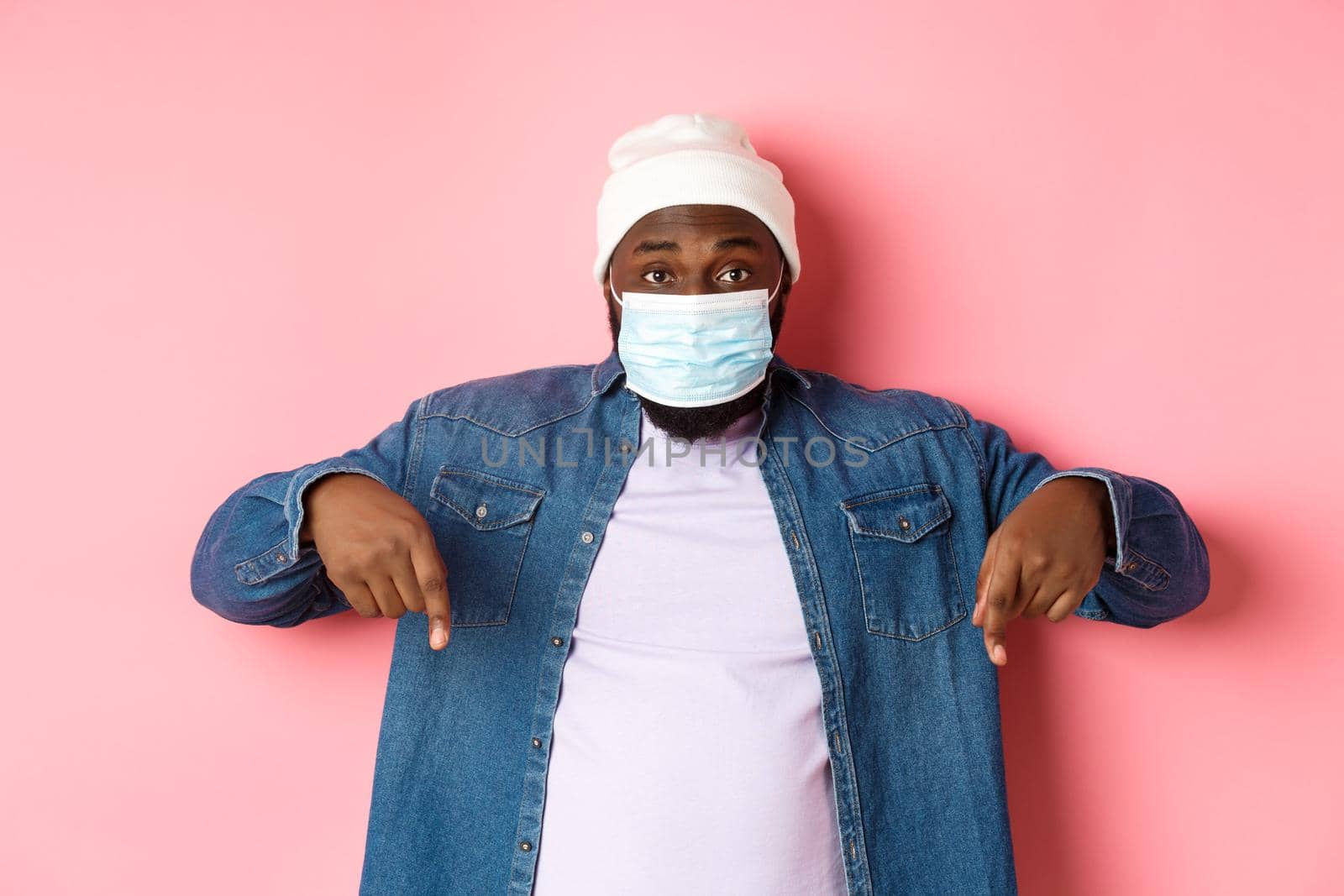 Covid-19, lifestyle and lockdown concept. Handsome african-american man in face mask, pointing fingers down, showing promo offer on bottom, standing over pink background.