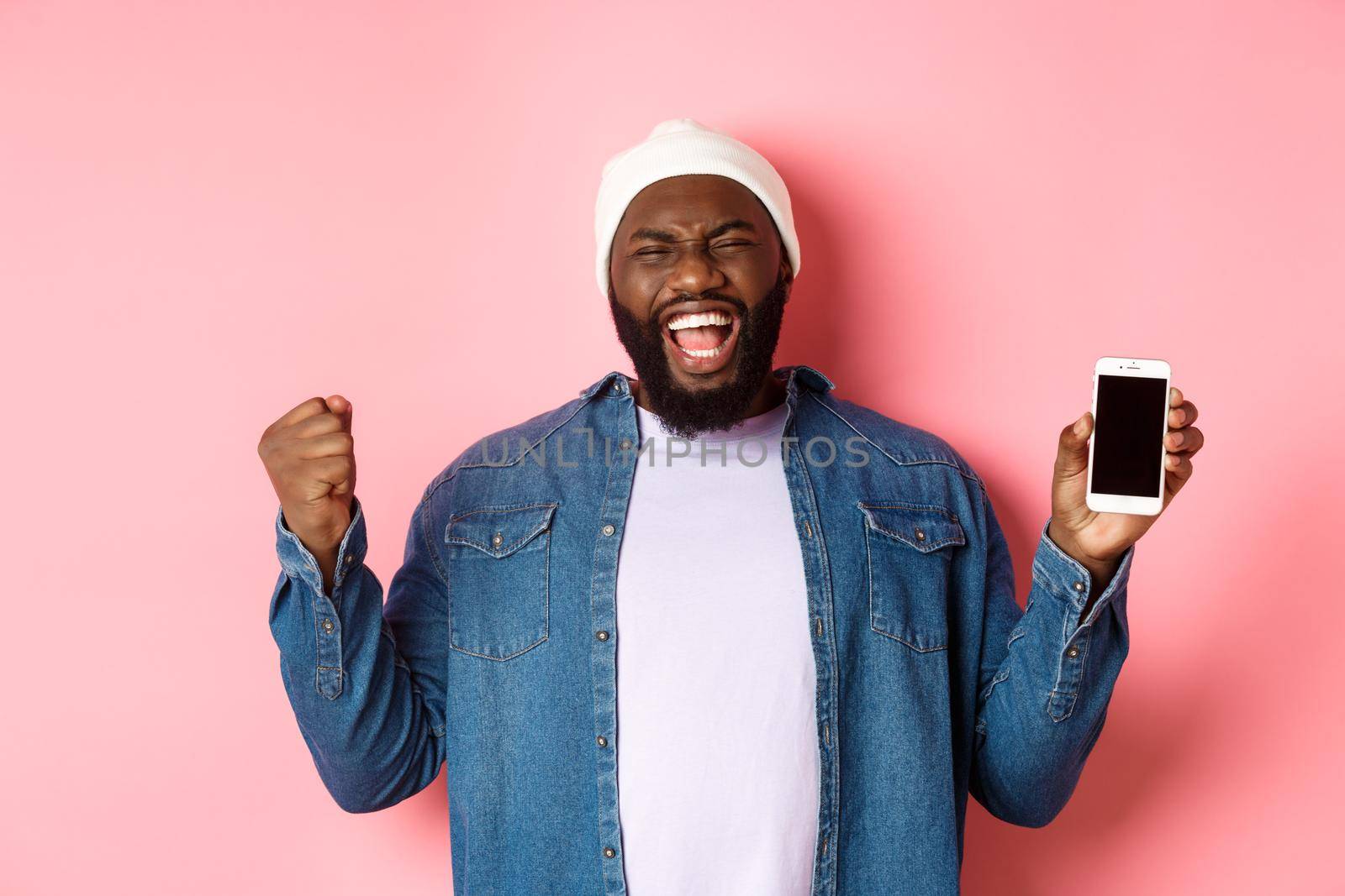 Happy Black guy celebrating online achievement, showing smartphone screen, rejoicing and scream yes with satisfaction, standing over pink background.