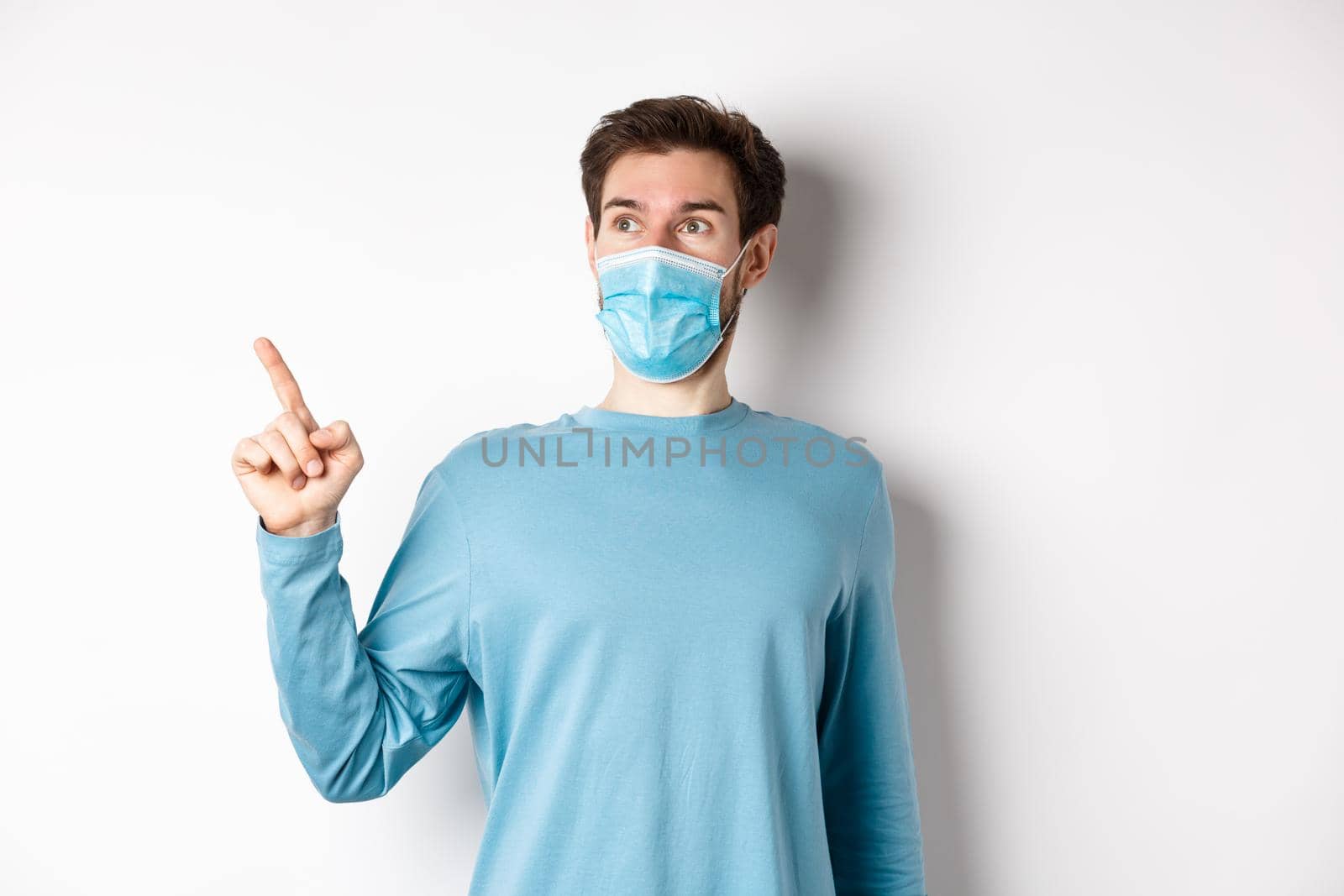 Coronavirus, health and quarantine concept. Surprised guy looking in awe and pointing at upper left corner, standing in medical mask and sweatshirt over white background.