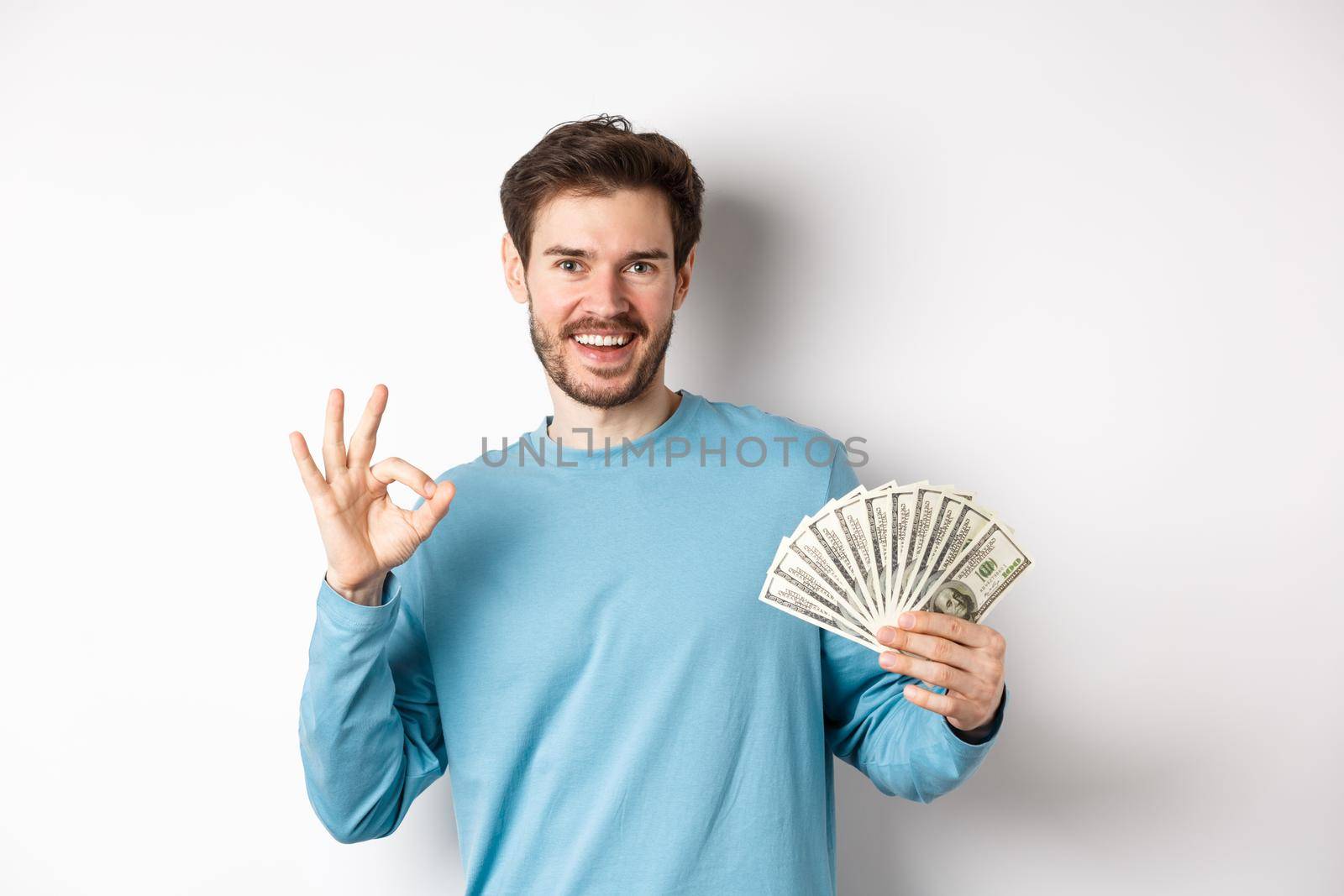 Handsome young man showing quick loans money, make okay gesture and smiling with cash, standing over white background by Benzoix