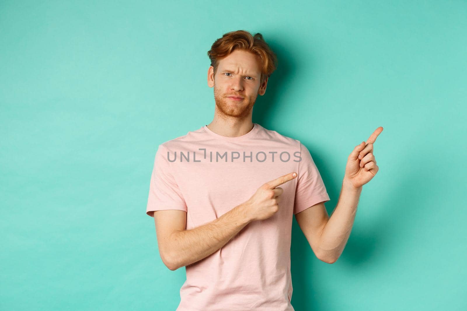 Skeptical and doubtful redhead man in t-shirt pointing fingers at upper right corner, showing promo offer with displeased face, standing over turquoise background by Benzoix