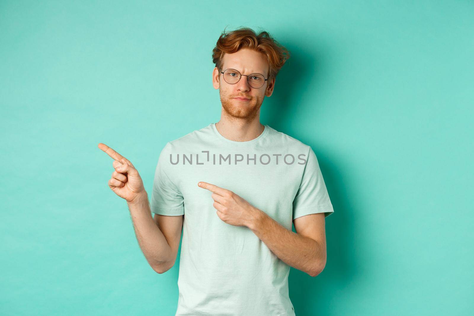 Judgemental redhead man in glasses grimacing, frowning disappointed and pointing left, showing bad promo offer, standing over turquoise background by Benzoix