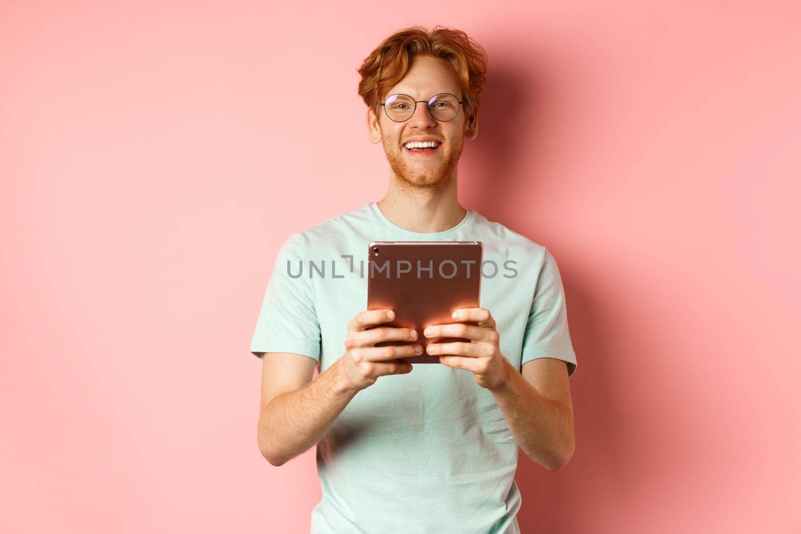 Cheerful caucasian man in t-shirt and glasses using digital tablet, looking happy at camera and smiling, standing over pink background.