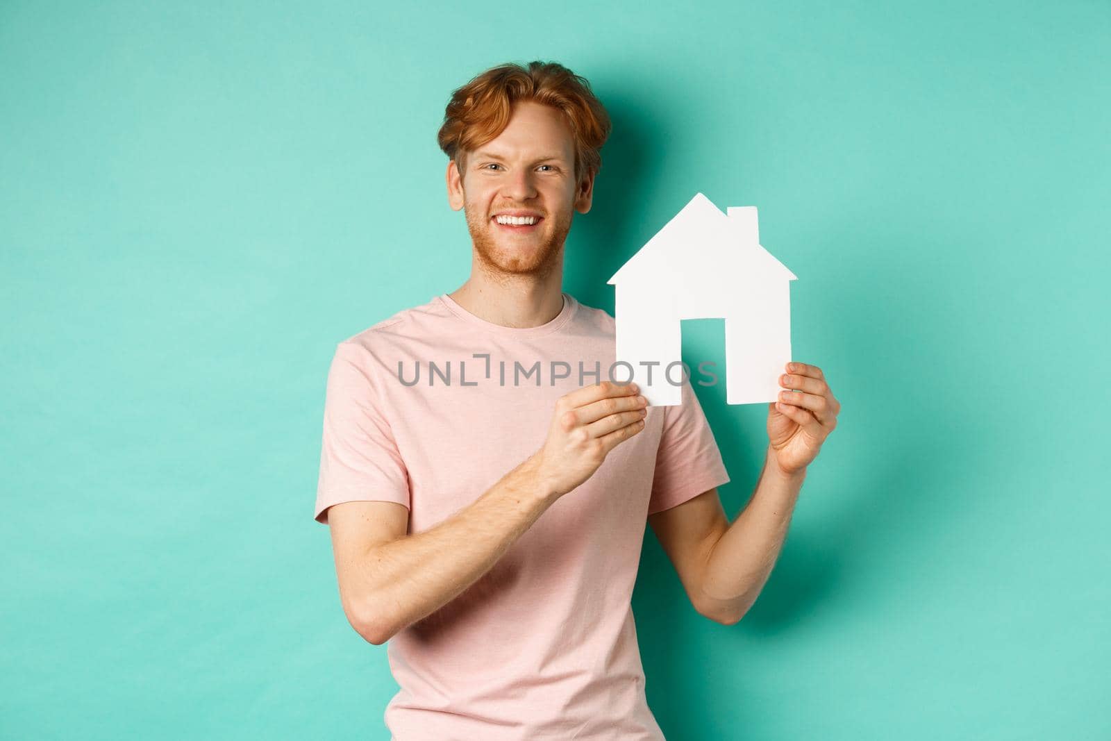 Real estate concept. Young man with red hair, wearing t-shirt, showing paper house cutout and smiling happy, standing over mint background.