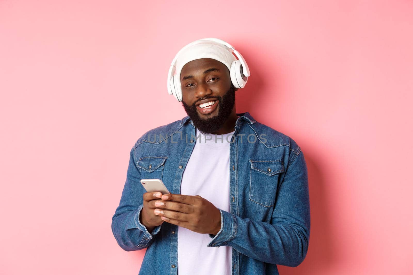 Handsome hipster guy in headphones smiling satisfied at camera, listening music in headphones, using mobile app, standing over pink background by Benzoix