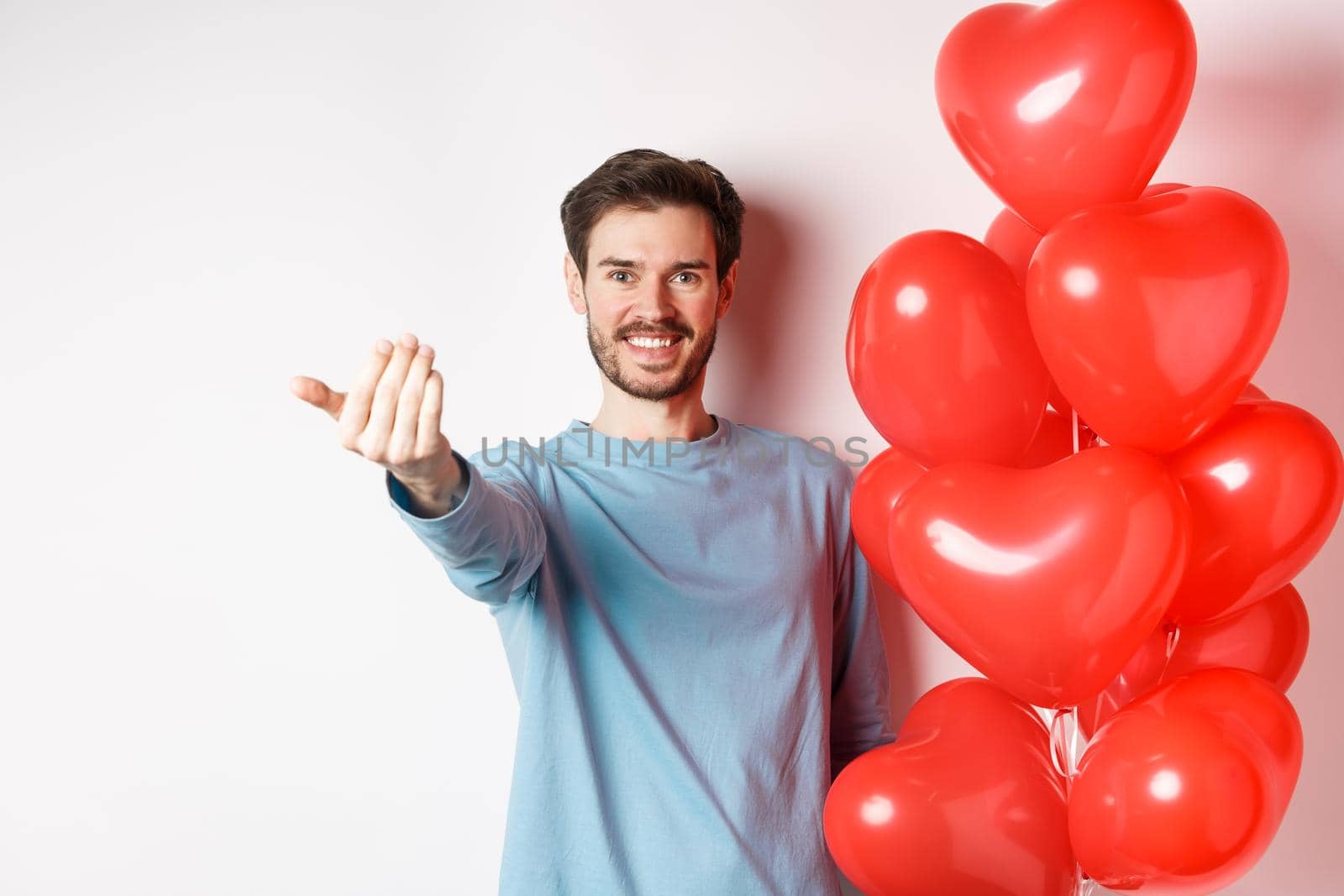 Smiling man beckon you to come closer, follow me gesture, taunting his lover move forward, have romantic surprise, standing near red balloon on valentines day, white background.