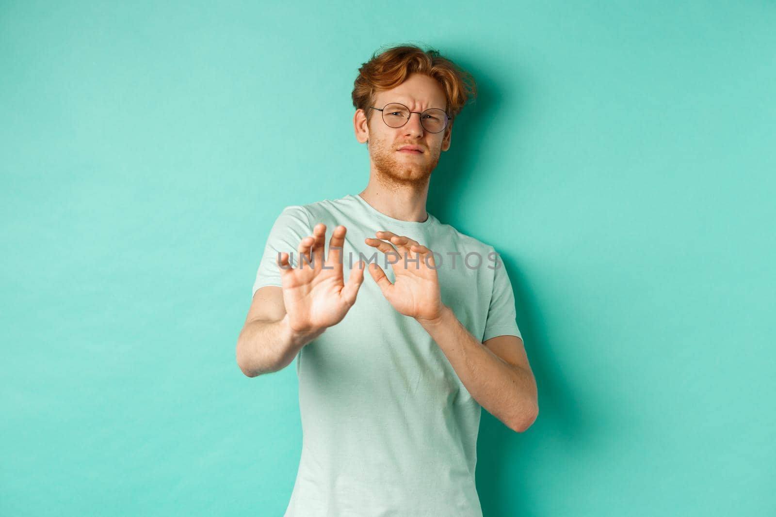 Reluctant redhead man in glasses raising hands in refusal gesture, step backwards, avoiding something with disgusted and unamused face, standing over turquoise background by Benzoix
