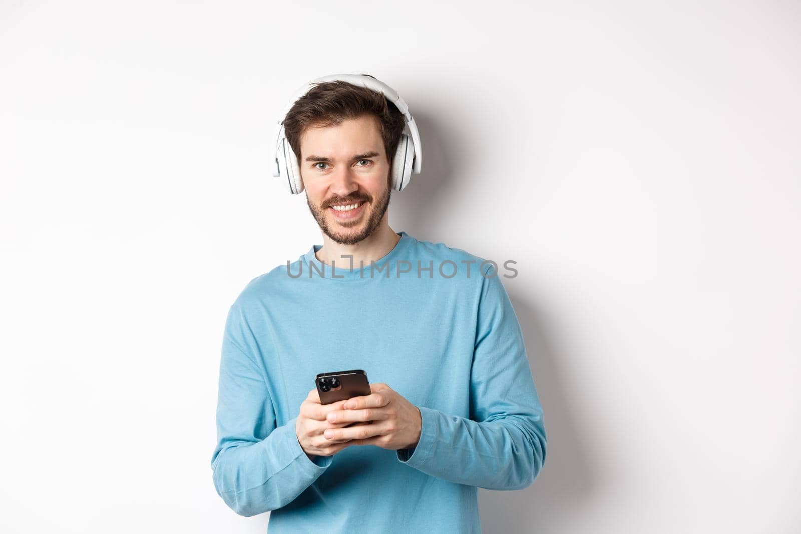 Attractive smiling man listening music in wireless headphones, using black smartphone and looking pleased, white background by Benzoix