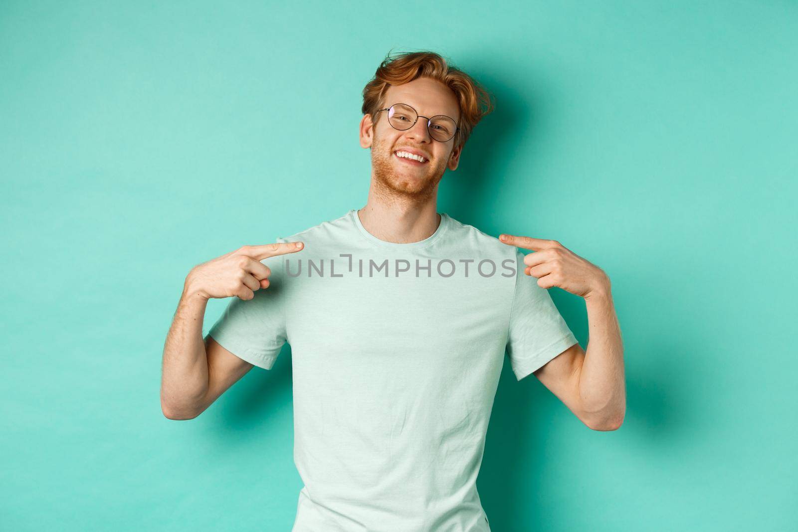 Confident redhead man in glasses and t-shirt, smiling with smug face and pointing at himself, bragging while standing over turquoise background.