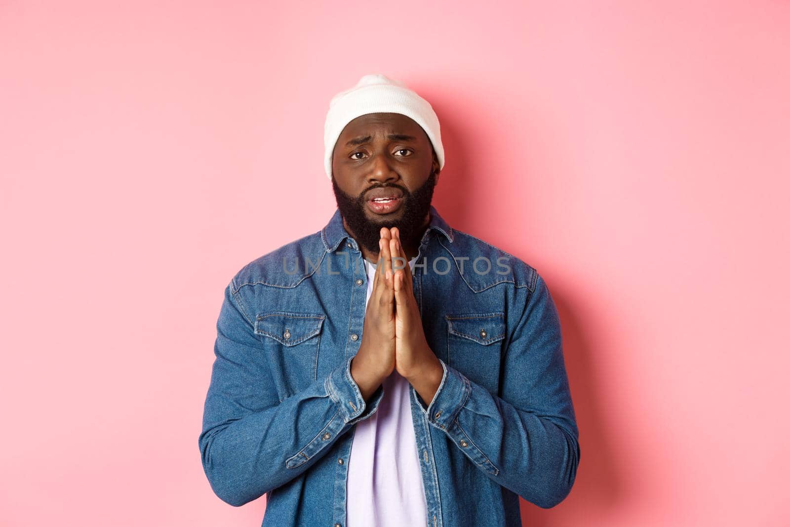 Upset african-american man asking for help, holding hands in pray, staring at camera sad and pleading, standing over pink background.