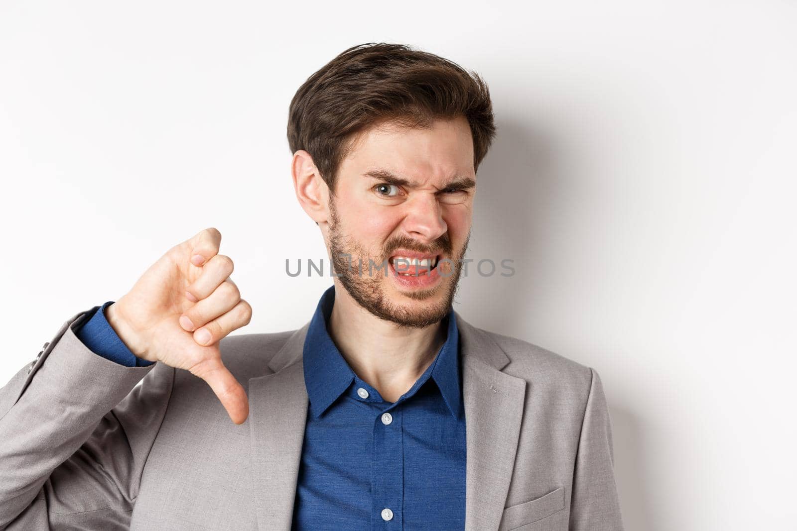 Close-up of disgusted guy in suit grimacing upset, showing thumbs down, express negative emotion, white background.