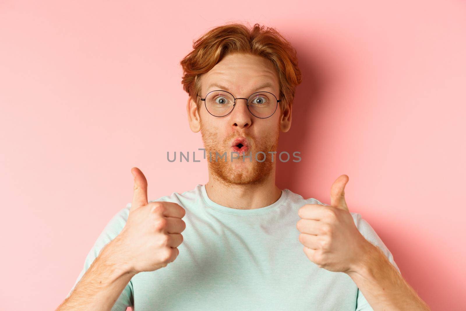 Face of happy redhead man in glasses and t-shirt, showing thumbs-up and looking excited, approve and praise cool promotion, standing over pink background.