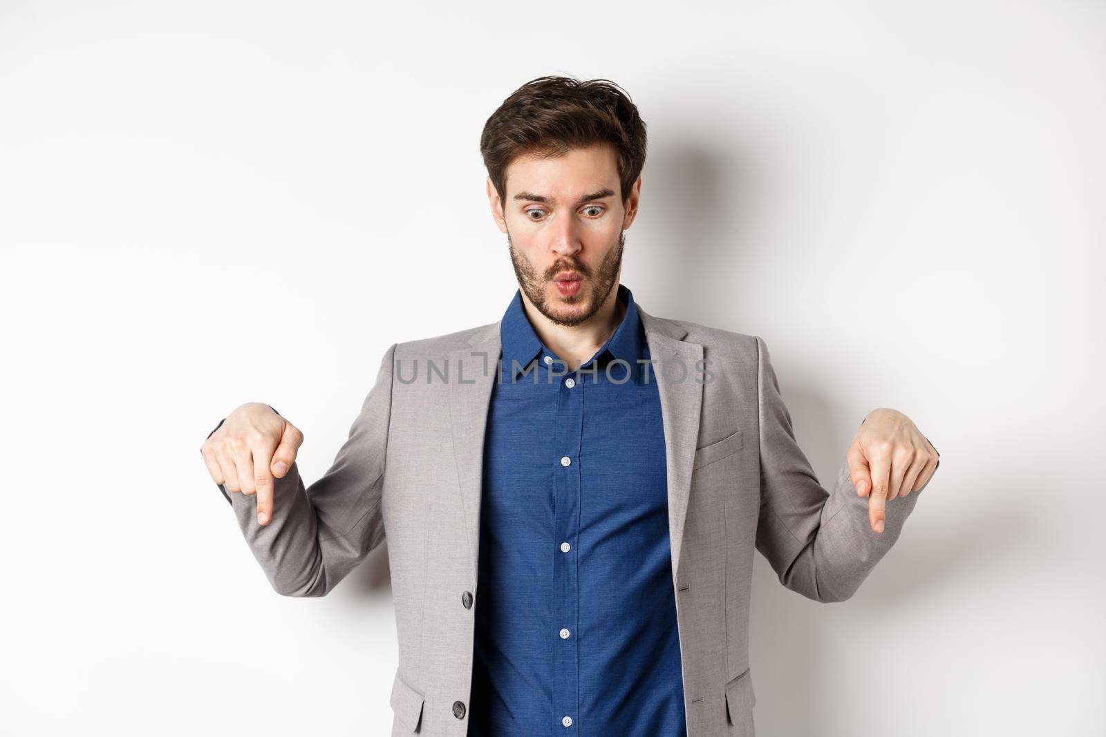 Wow look at this. Impressed and excited businessman in suit pointing fingers down, looking at bottom advertisement with amazed face, standing on white background by Benzoix