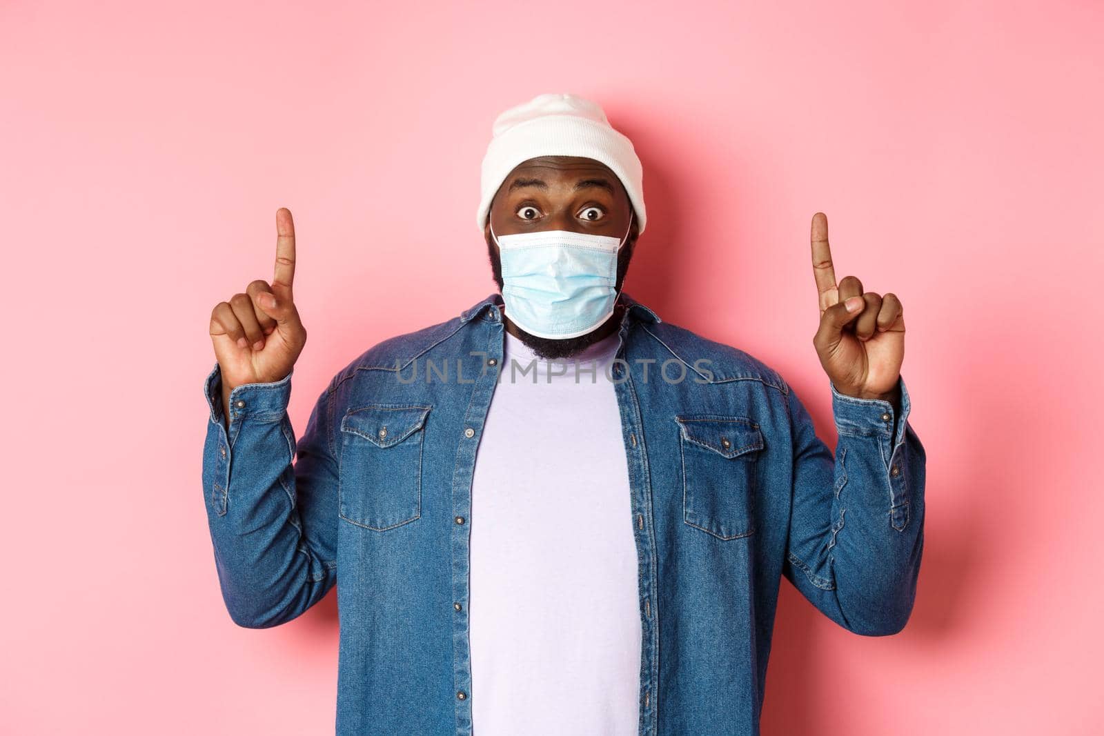 Coronavirus, lifestyle and global pandemic concept. Excited african-american guy in medical mask showing announcement, pointing fingers up, standing over pink background.