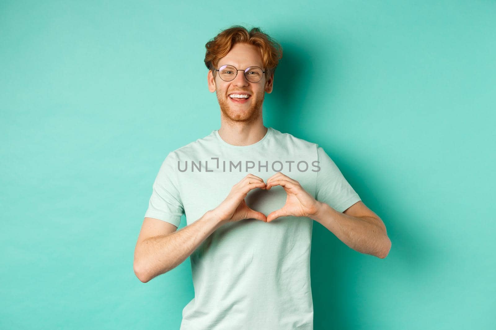 Valentines day and relationship concept. Happy boyfriend with red hair and beard, wearing glasses and t-shirt, showing heart sign and saying I love you, standing over turquoise background by Benzoix