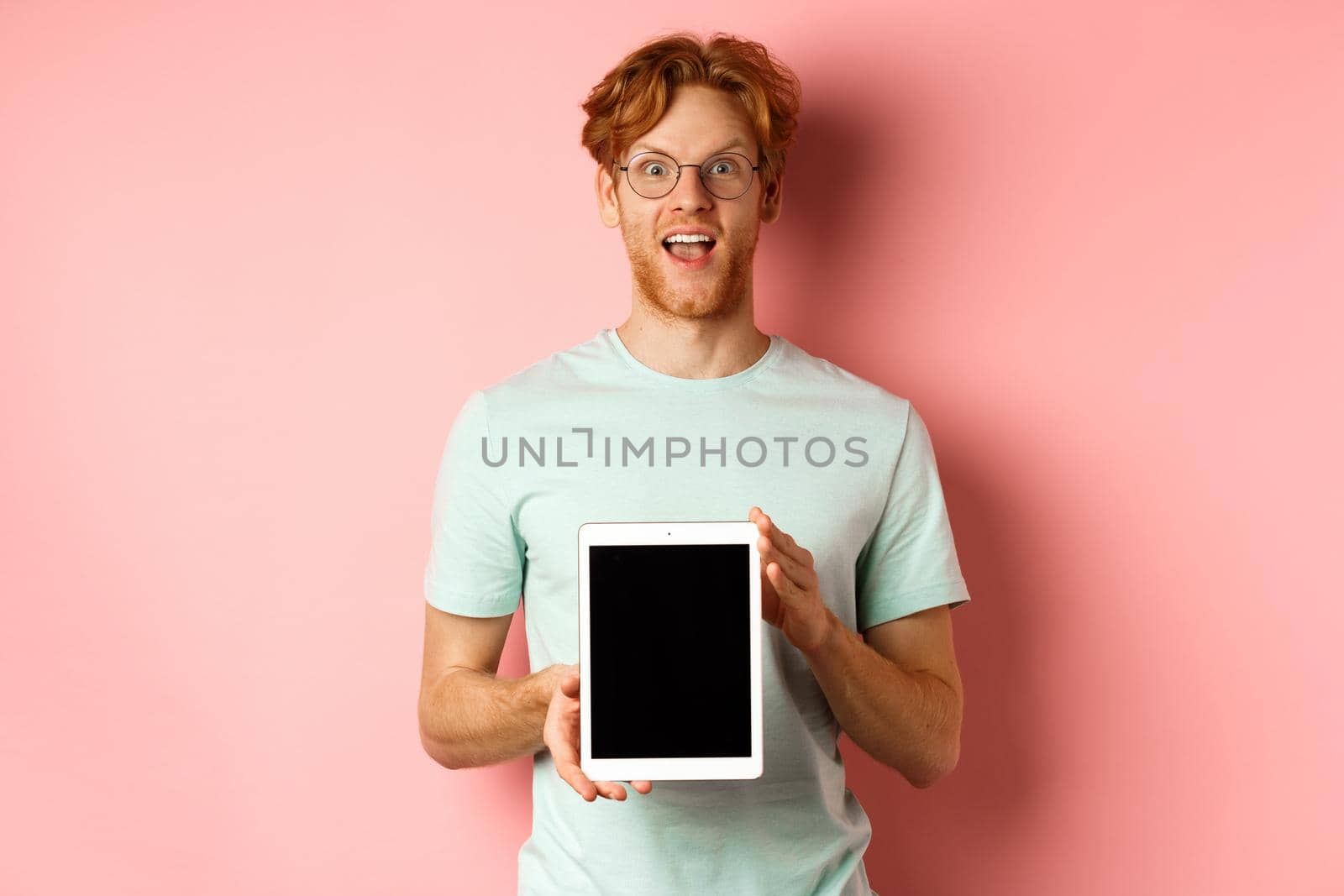 Excited young man with red hair and beard, checking out online promotion, showing digital tablet screen and staring at camera amazed, standing over pink background.