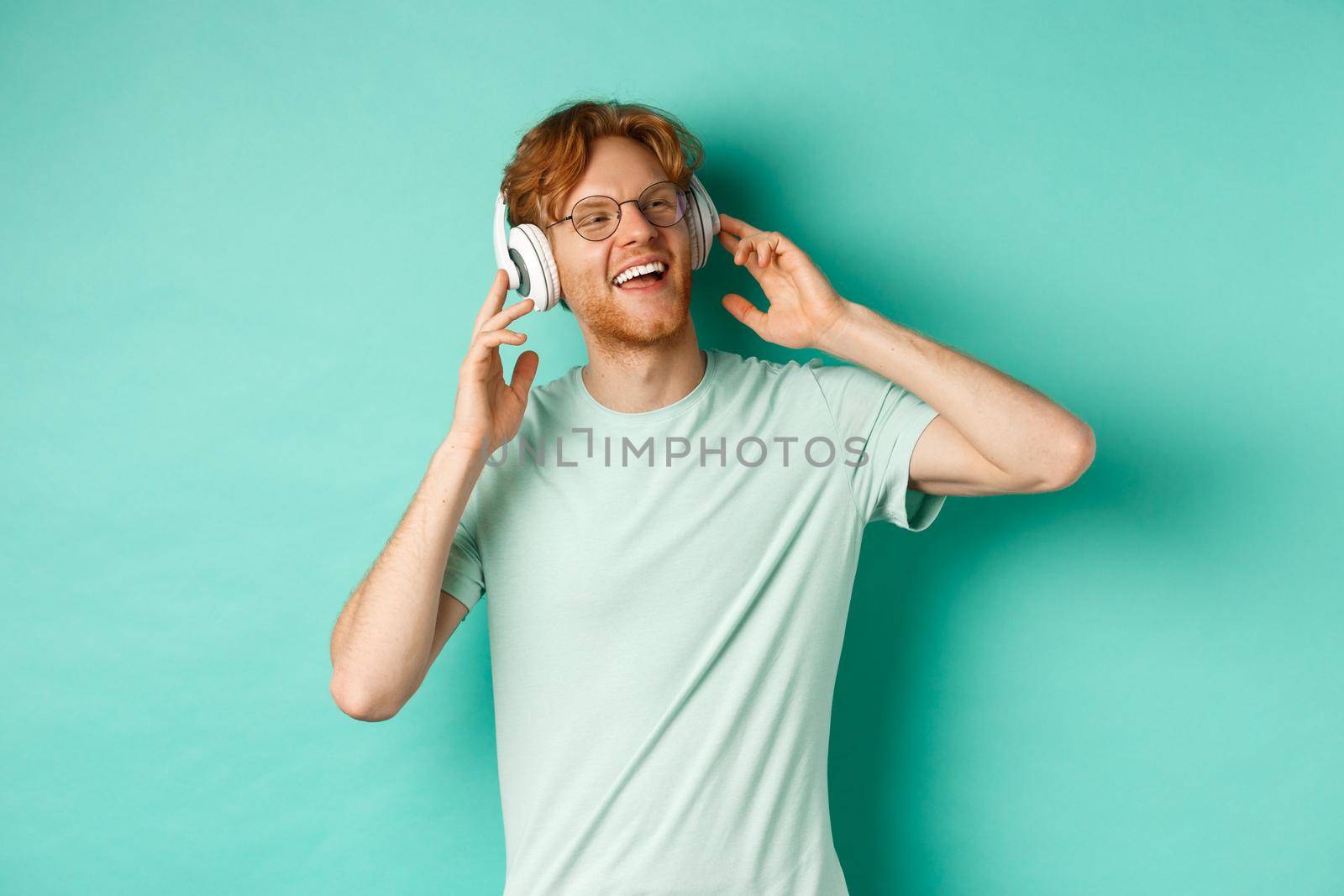 Lifestyle concept. Happy young man with ginger hair dancing and having fun, listening music on wireless headphones and smiling pleased, turquoise background.