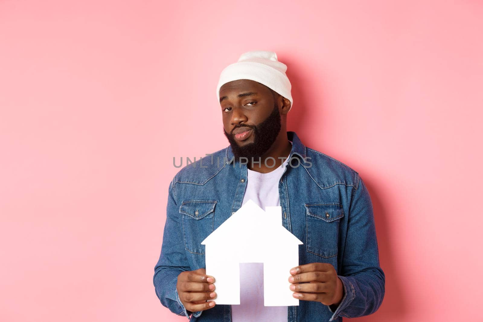 Real estate concept. Skeptical Black male model holding paper house model, looking suspicious and doubtful at camera, searching apartment, standing over pink background by Benzoix