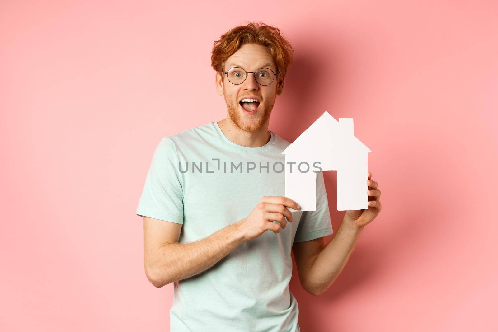 Real estate. Cheerful redhead man buying property, showing paper house cutout and smiling amazed, standing over pink background.