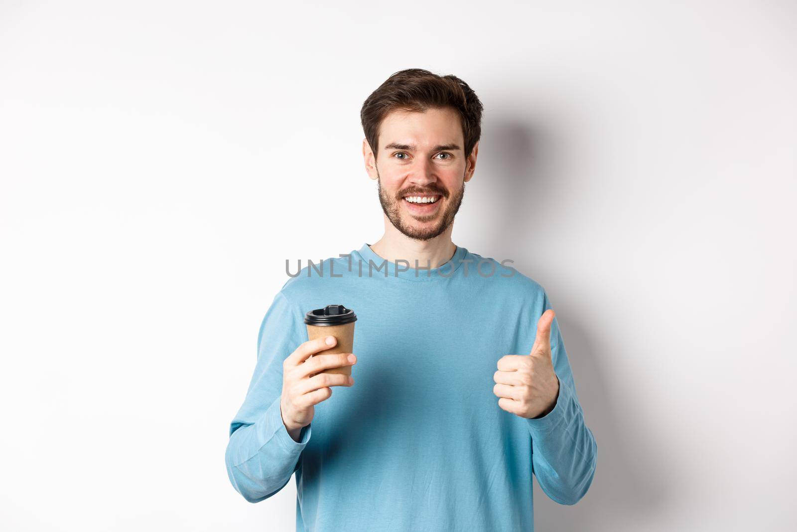 Cheerful young man drinking good coffee, holding paper cup and showing thumb up, recommending cafe shop, standing over white background by Benzoix