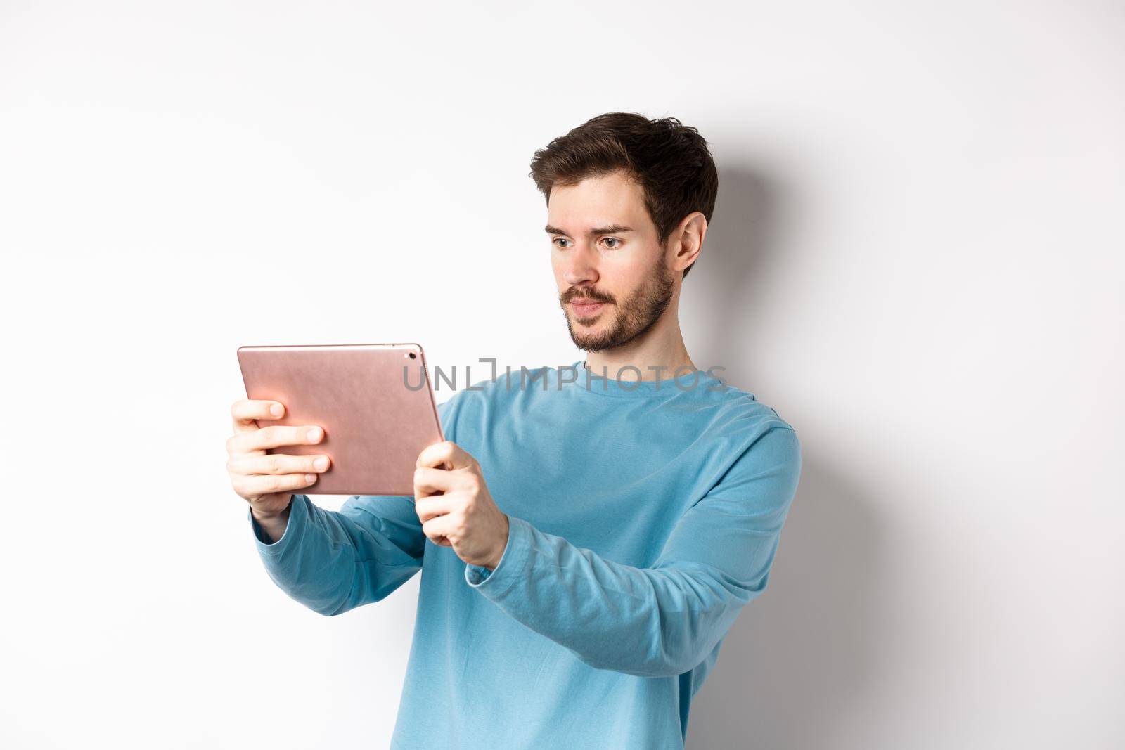 Young man taking photo on digital tablet, looking serious at gadget screen, standing over white background by Benzoix