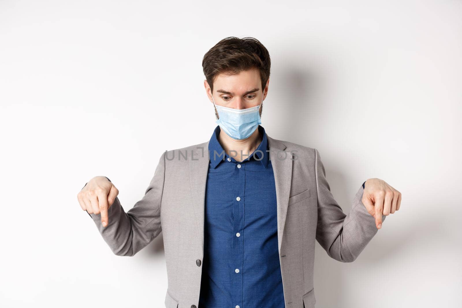 Covid-19, pandemic and business concept. Surprised guy in medical mask and suit looking and pointing down with popped eyes, gasping amazed, white background.