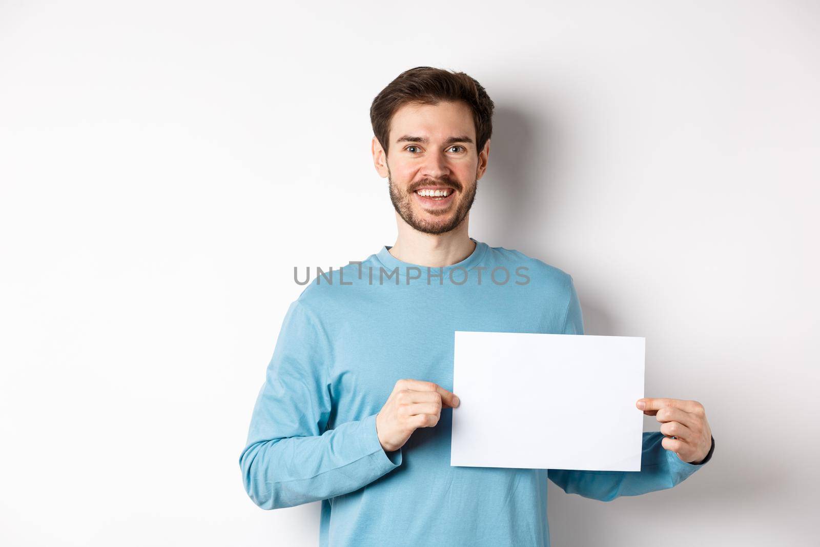 Young man with happy face showing blank piece of paper for your logo or sign, smiling at camera, standing over white background by Benzoix