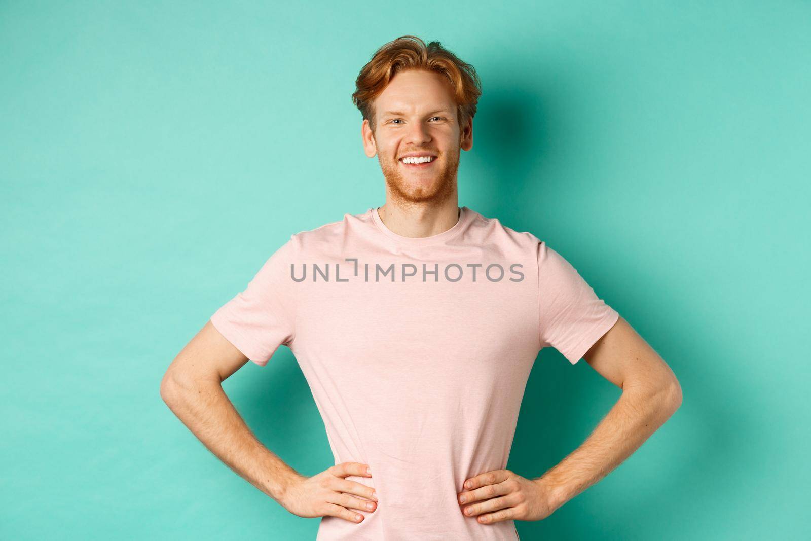 Enthusiastic young man with red hair, wearing t-shirt, standing happy and proud with hands on hops, standing over turquoise background.