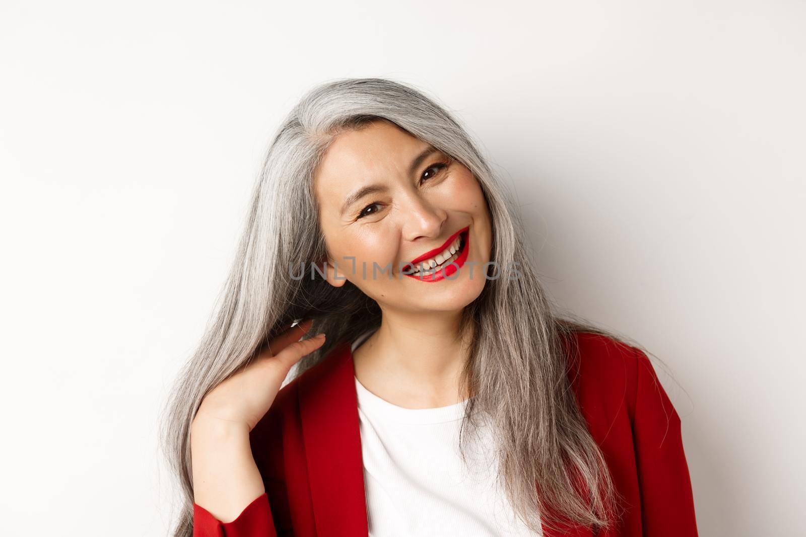 Beauty and aging concept. Close up of asian senior woman with red lips, long healthy grey hair, smiling at camera, standing over white background.