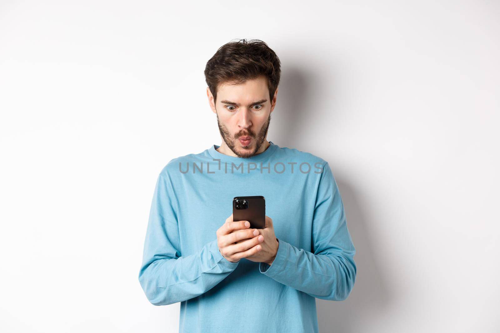 Amazed young man staring at smartphone screen and saying wow, looking at online offer, standing on white background by Benzoix