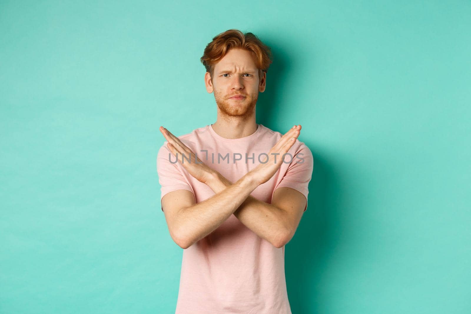 Skeptical redhead man in t-shirt saying no, cross arms on chest and frowning disappointed, prohibit something bad, standing over turquoise background.