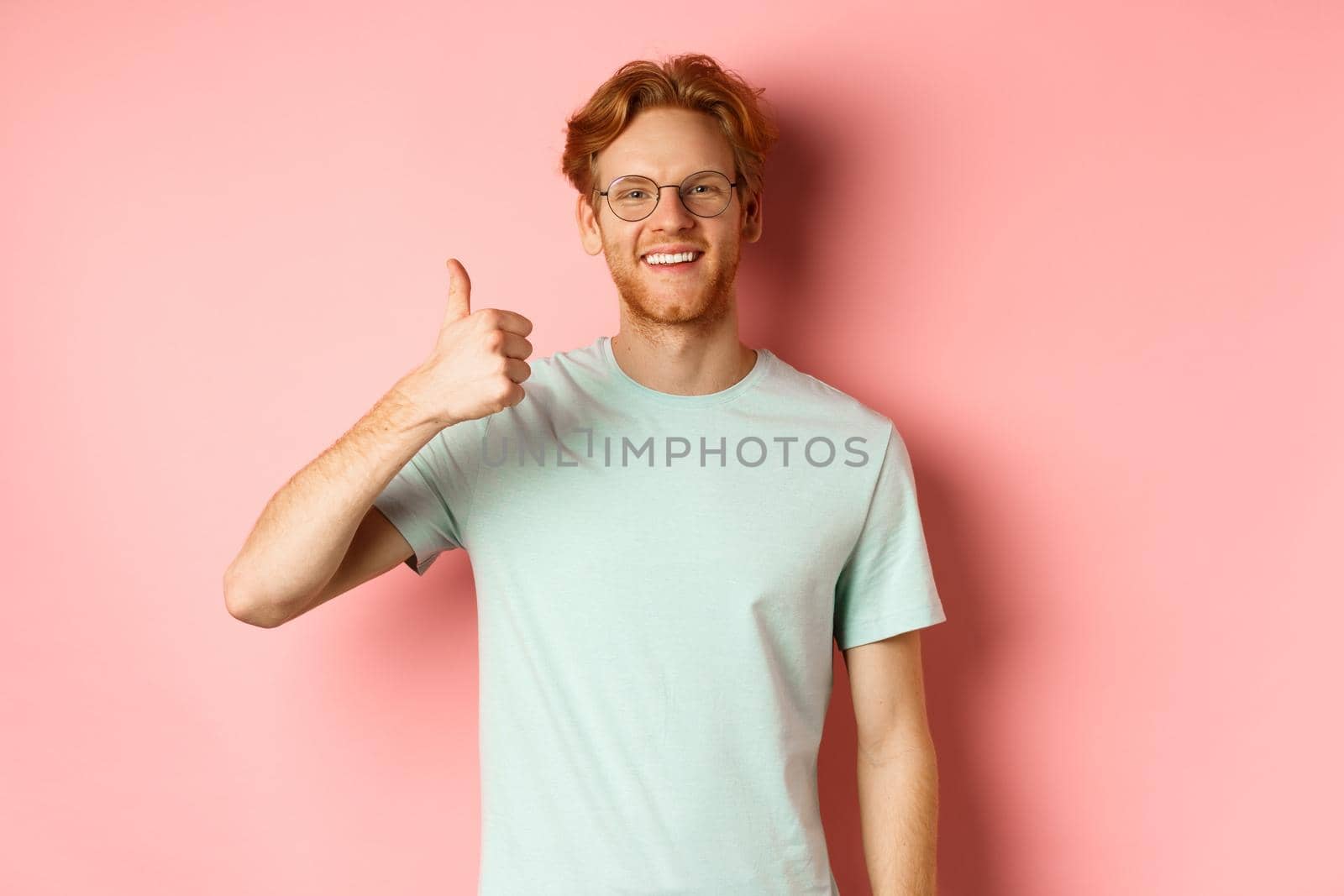 Cheerful redhead man in glasses and t-shirt, showing thumbs up with satisfied face, showing positive reaction, approve and agree with you, standing over pink background.