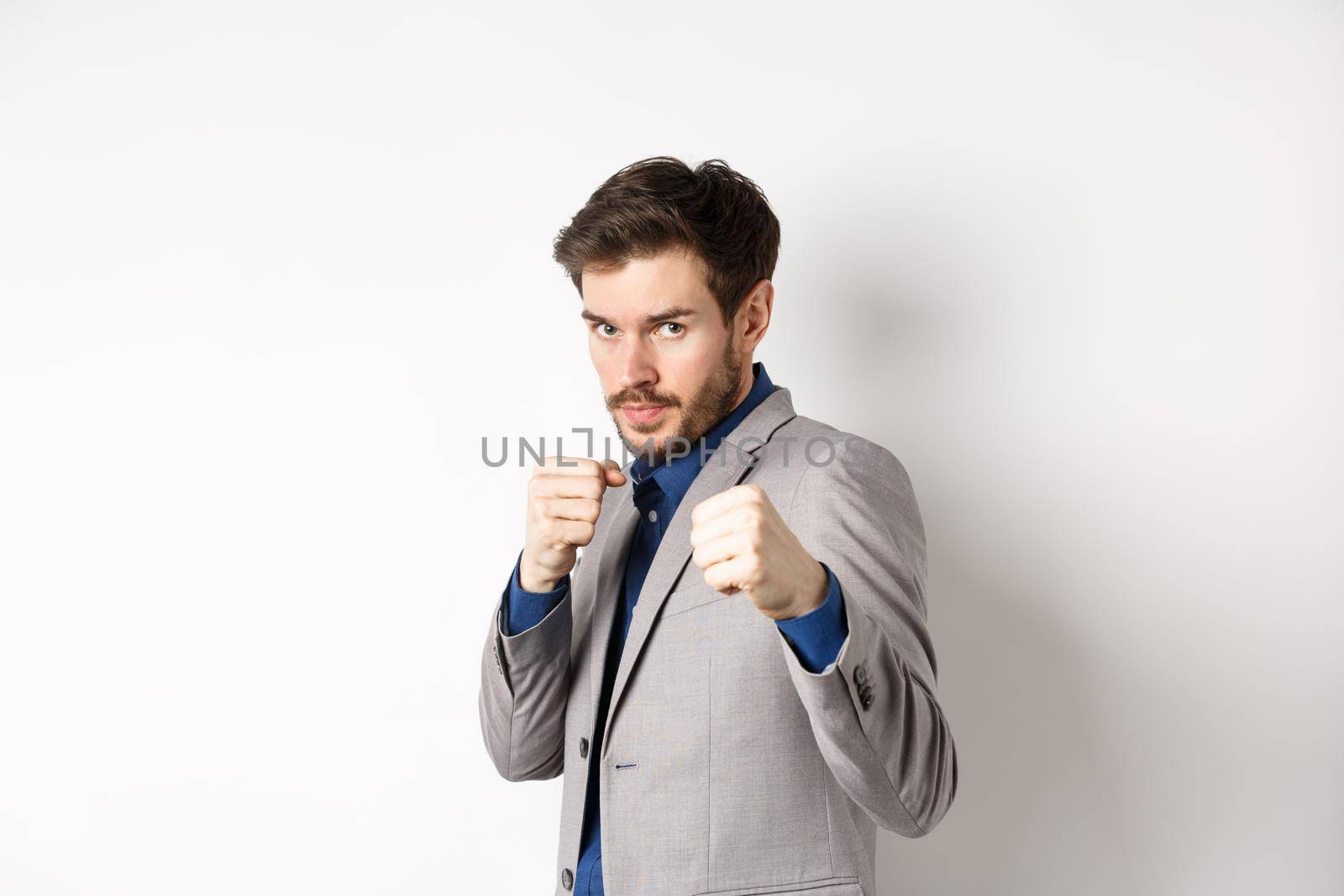 Serious man in suit raising hands in boxer pose, going to fight, looking confident, beckon to attack, standing on white background in defensive pose.
