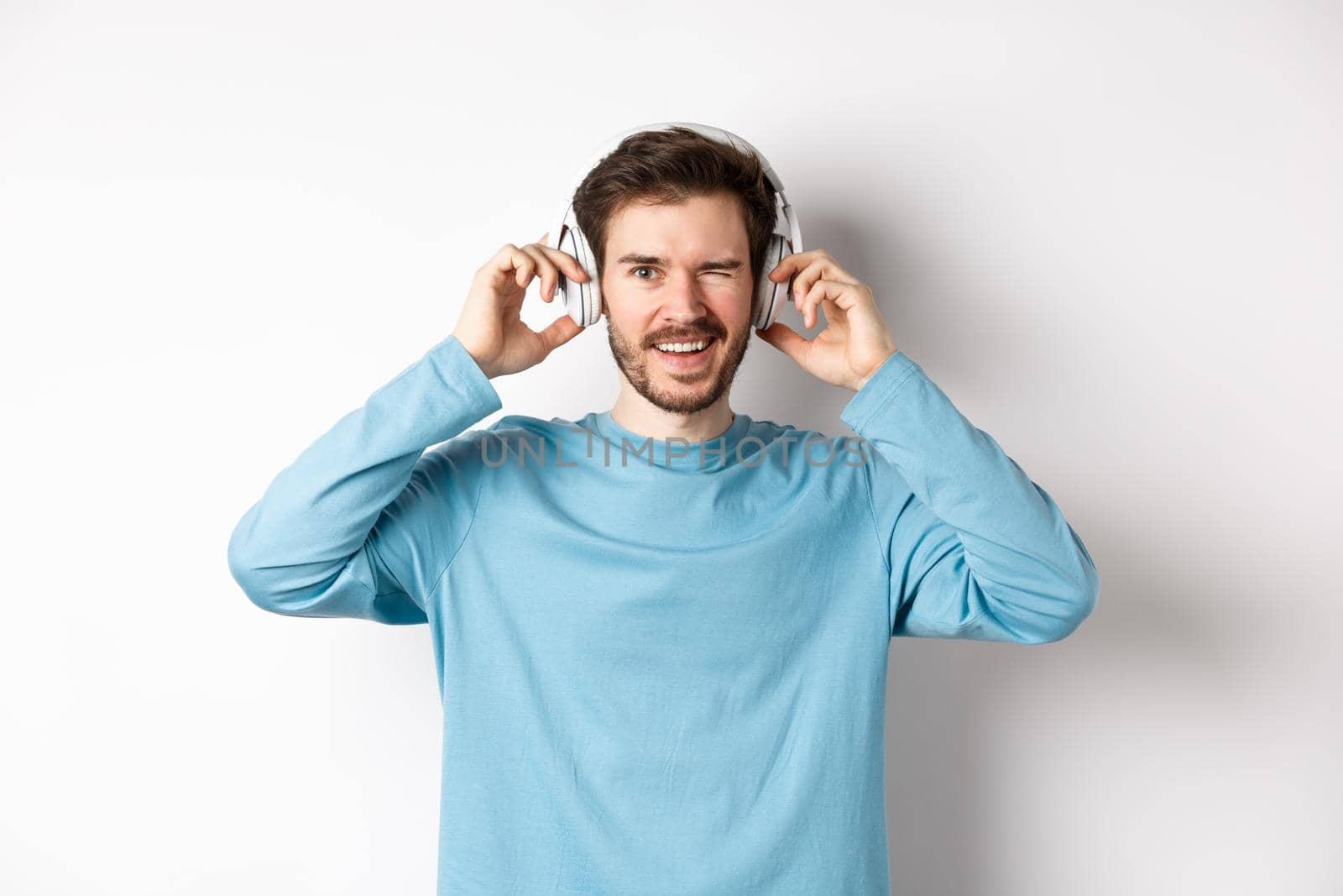 Cheerful guy wink at camera and smiling, listening music in wireless headphones, white background by Benzoix
