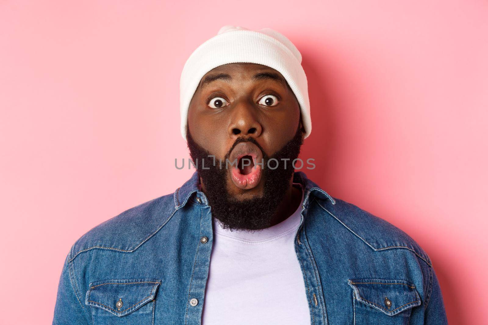 Close-up of impressed Black male model saying wow, staring at camera amazed and wondered, standing over pink background by Benzoix