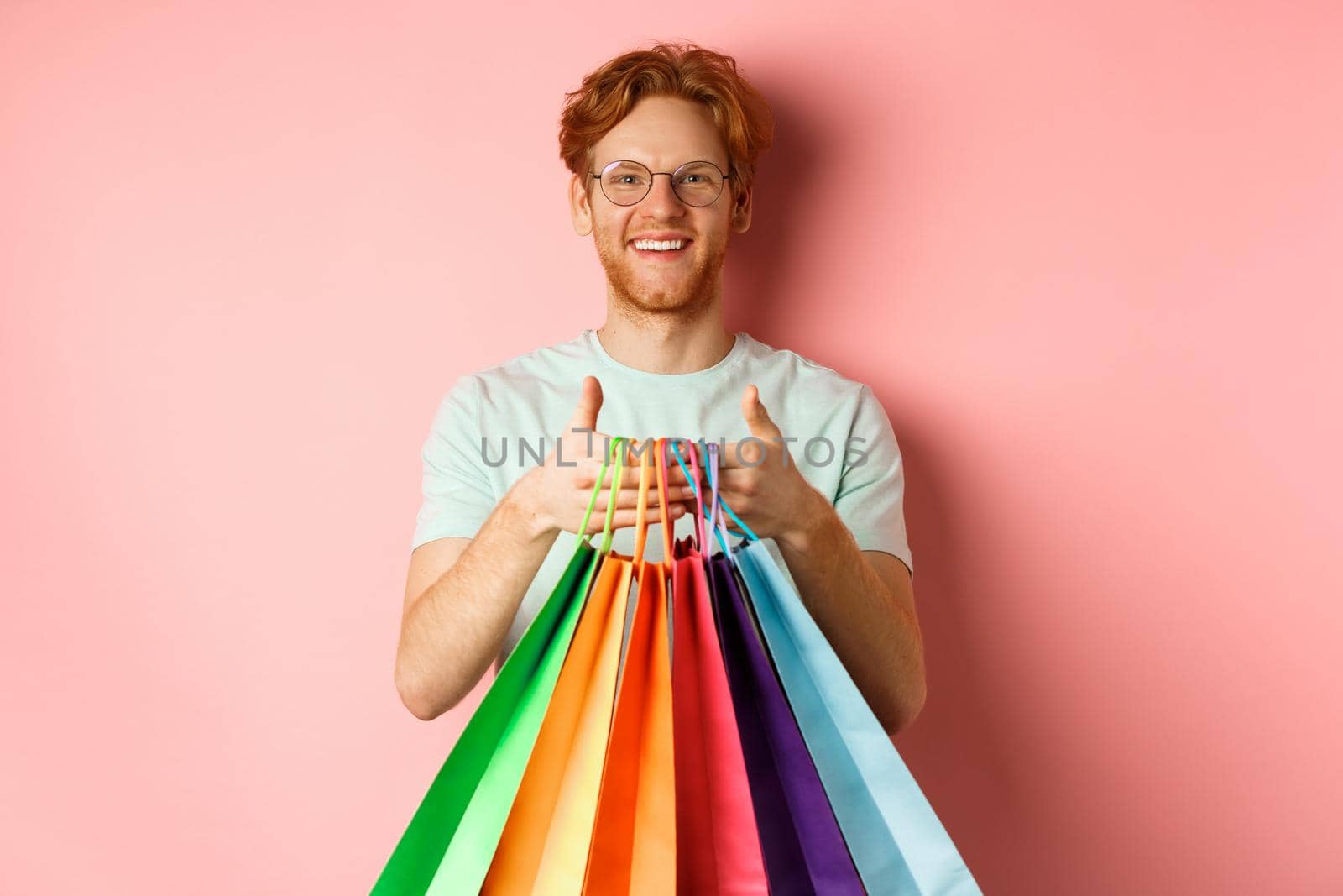 Cheerful redhead man buying gifts, holding shopping bags and smiling, standing over pink background.