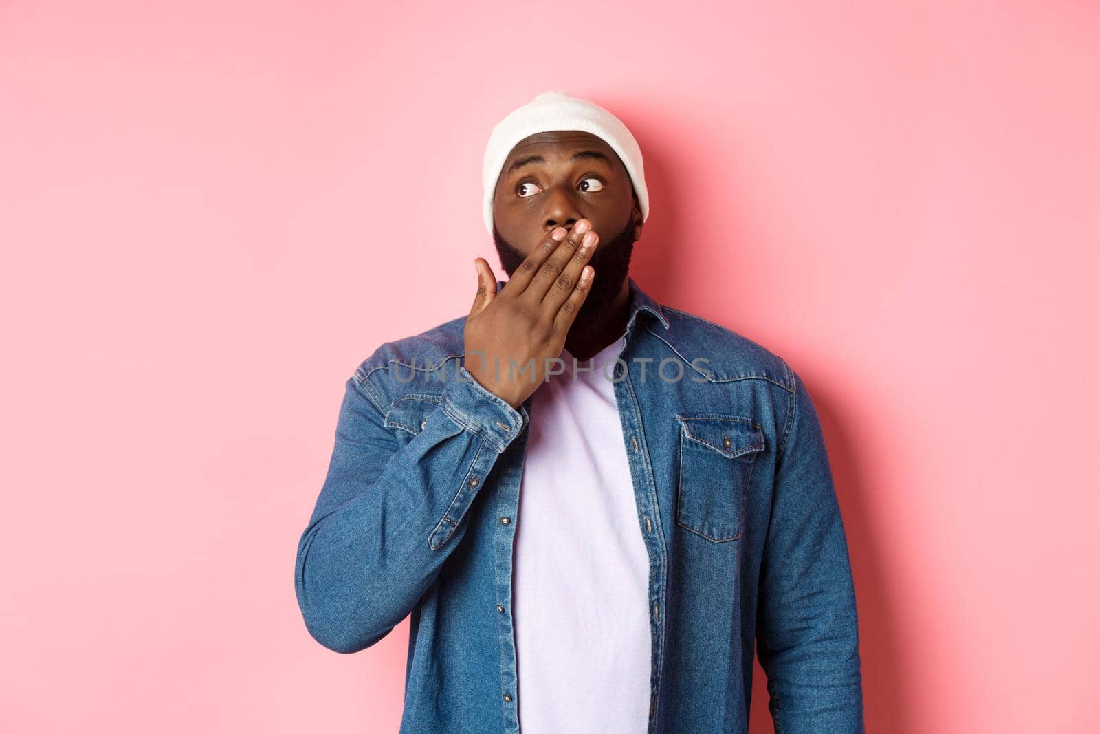 Shocked african-american man gasping, staring left in awe, gossiping, standing over pink background.