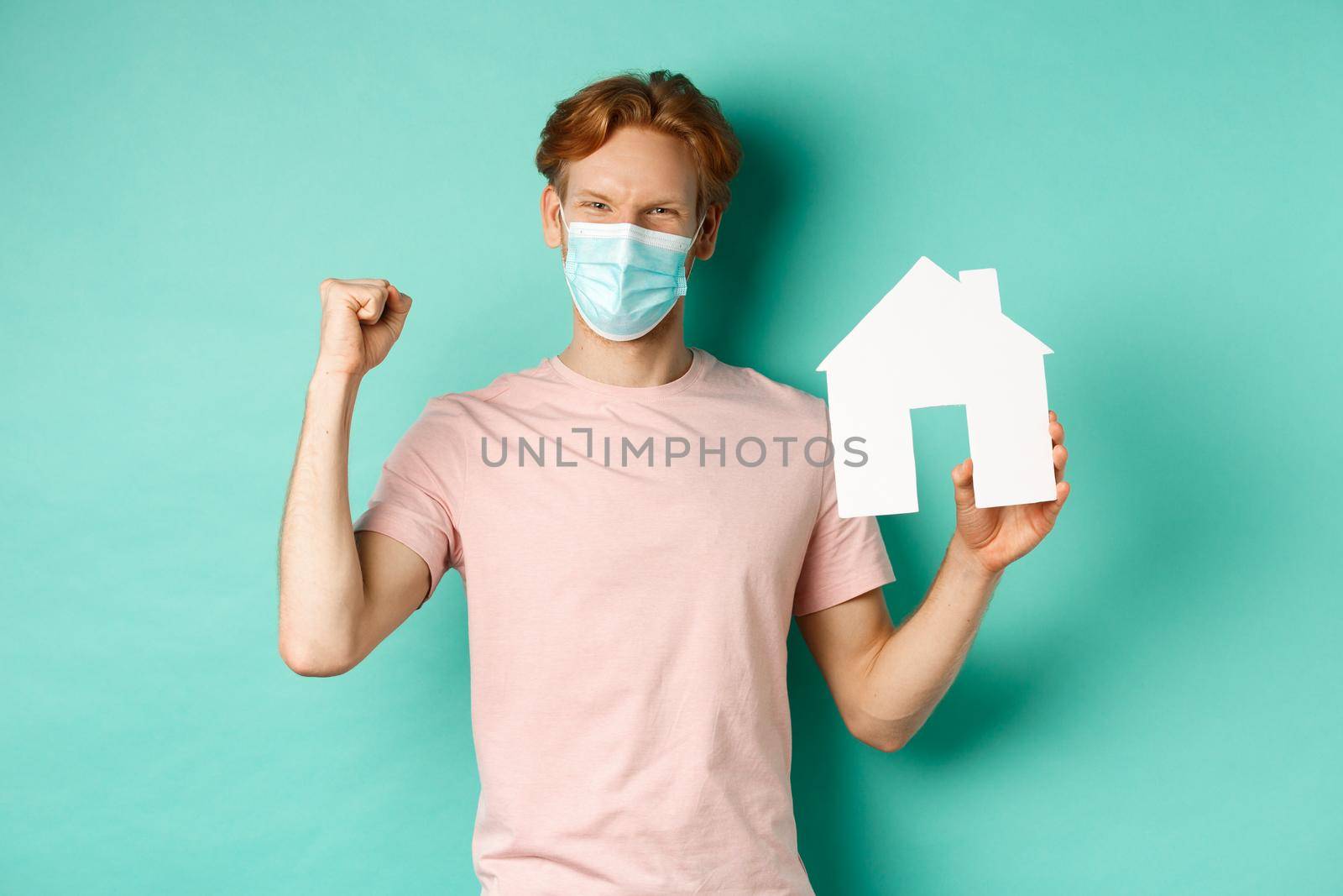Covid-19 and real estate concept. Happy redhead man in medical mask, showing paper house cutout and fist pump, rejoicing and winning, standing over turquoise background.