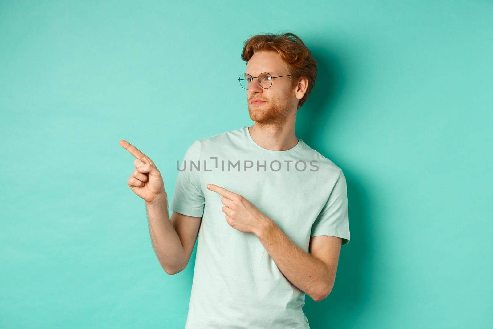Skeptical caucasian guy with red hair and beard, wearing glasses and t-shirt, looking and pointing left disappointed, judging something bad, standing over mint background.