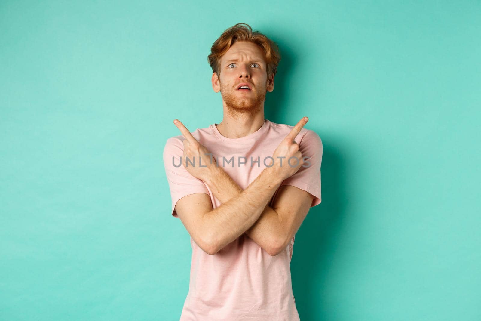 Confused young man in t-shirt looking up, pointing fingers sideways and feeling puzzled, cant decide, standing over turquoise background.