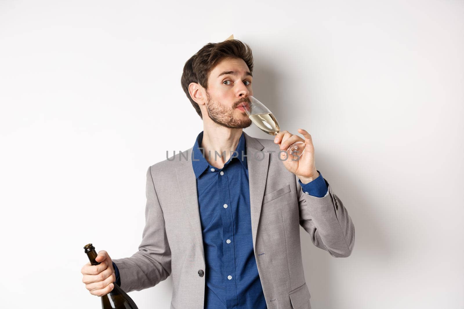 Celebration and holidays concept. Handsome bearded man in suit and birthday hat holding bottle, drinking glass of champagne, standing on white background.