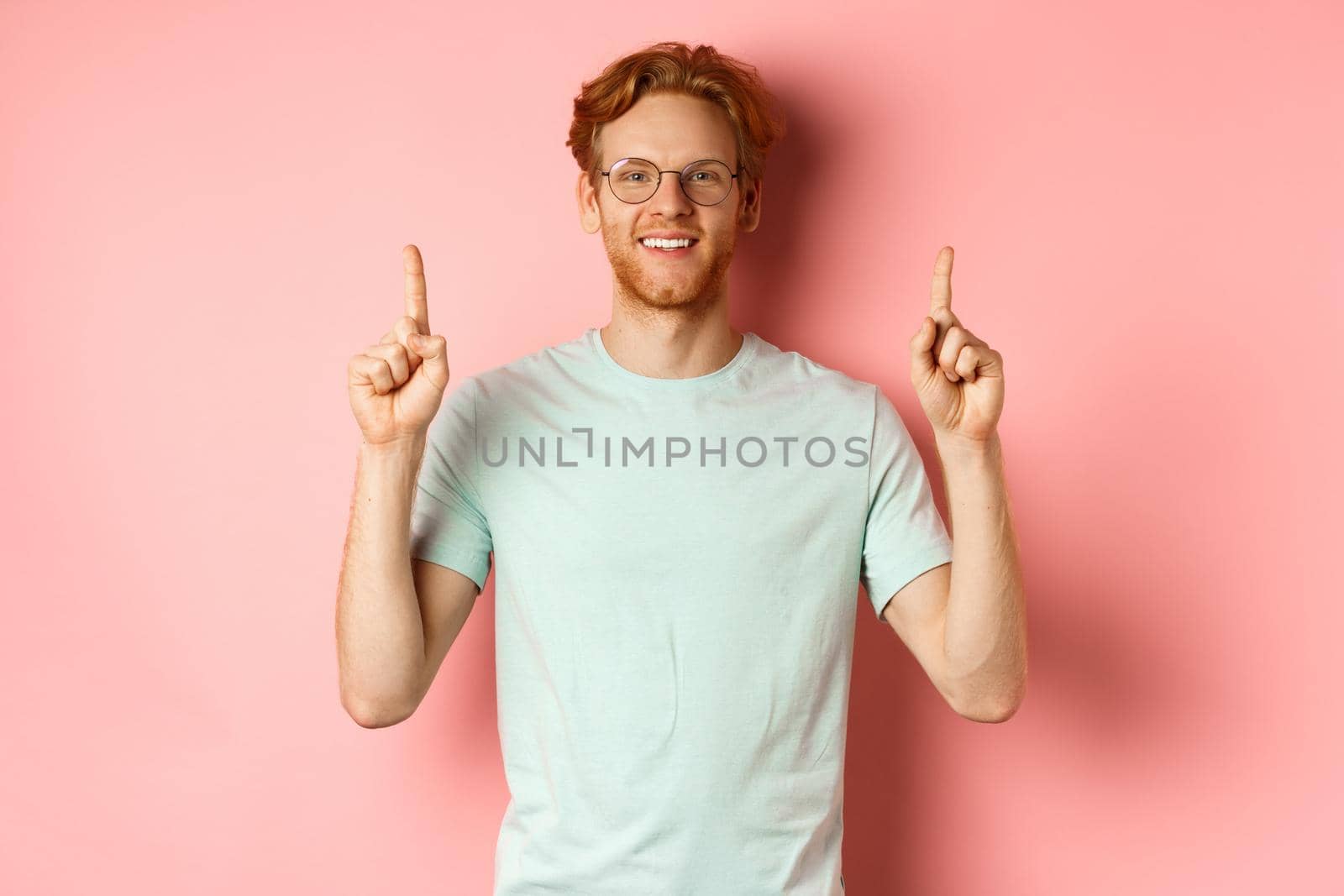 Attractive young man in glasses and t-shirt showing promo deal, pointing fingers up and smiling at camera, standing over pink background by Benzoix
