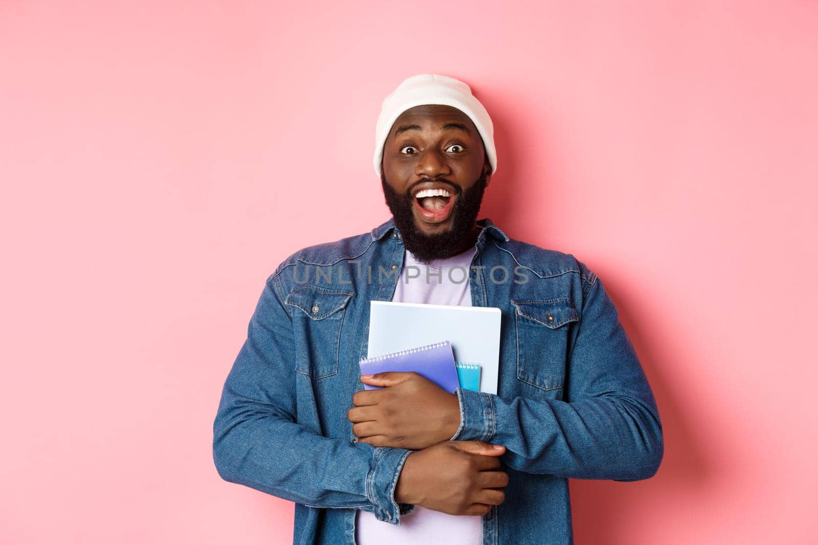 Image of adult african-american man holding notebooks and smiling, studying at courses, standing over pink background.