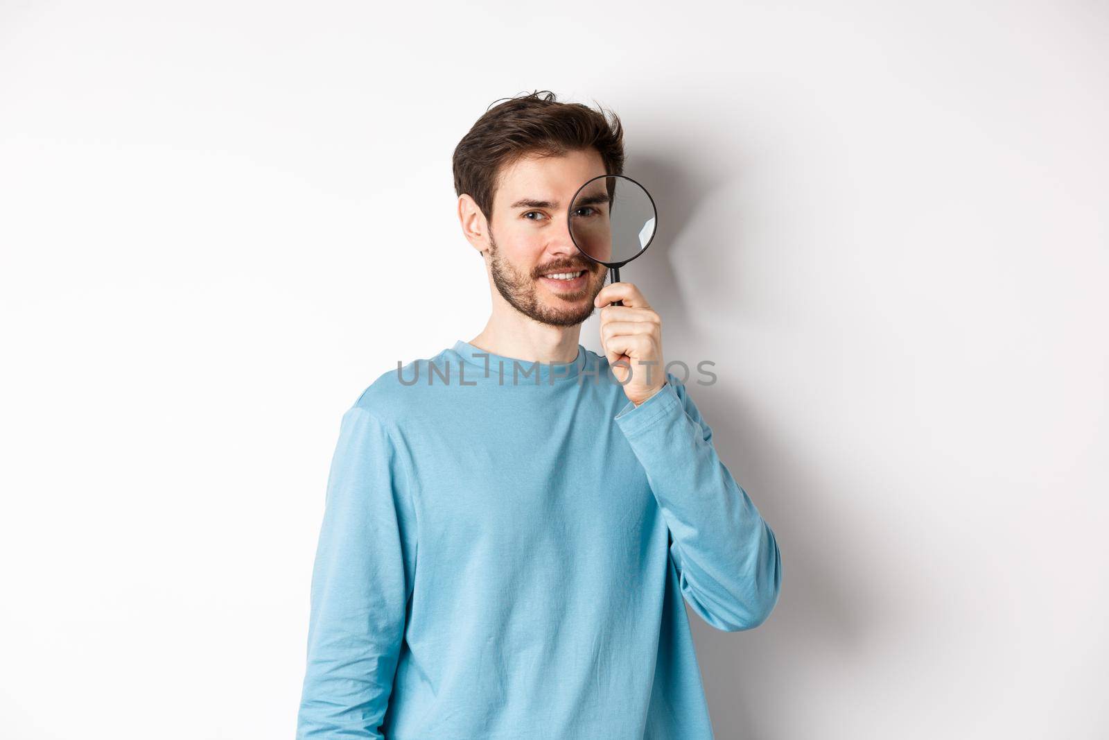 Smiling young man detective looking through magnifying glass, searching for promo offer, standing on white background.